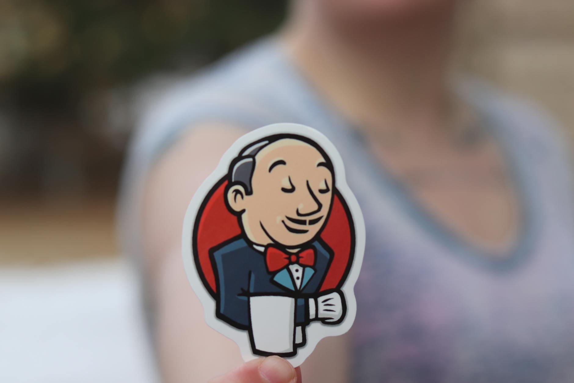 Pierwszy pipeline CI/CD: jak zautomatyzować testy i wdrożenia bez stresu Dłoń trzymająca naklejkę Jenkins na rozmytym tle na zewnątrz